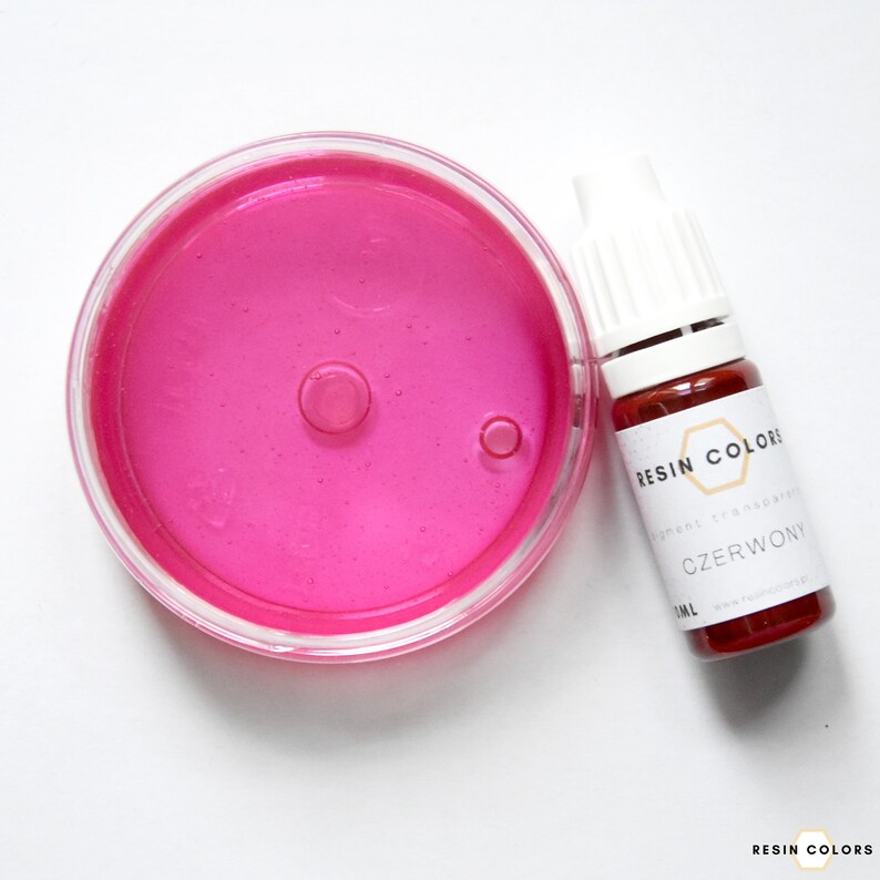 May include: A clear, round container filled with bright pink liquid next to a small bottle of red liquid. The bottle has a white cap and a label that reads "CZERWONY" and "RESIN COLORS".