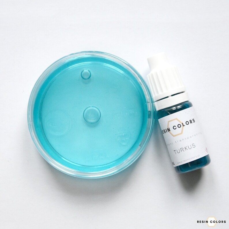 May include: A clear, round container with a light blue liquid and two raised circles. Next to it is a small bottle labeled "RESIN COLORS TURKUS" with a white cap. The liquid in the bottle is also light blue.