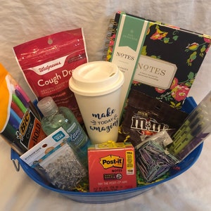 May include: A blue basket filled with a variety of gifts, including a coffee mug that says "make today amazing", a pack of M&Ms, a pack of Post-it notes, a bottle of hand sanitizer, a pack of push pins, a pack of Sharpie Clear View markers, a pack of BIC Crystal Bold pens, and two notebooks with floral designs.