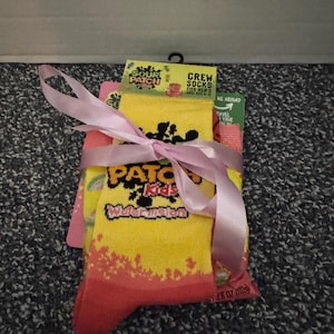 May include: A pair of yellow and pink crew socks featuring Sour Patch Kids branding and the text Watermelon, packaged with a matching bag of candy. The set is tied together with a light pink ribbon and includes a label stating fits shoe size 6-12.