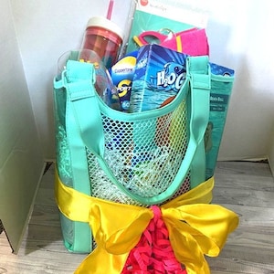 May include: A turquoise mesh beach bag with yellow ribbon and pink tissue paper. The bag is filled with beach essentials, including a water bottle, sunscreen, and a beach ball.