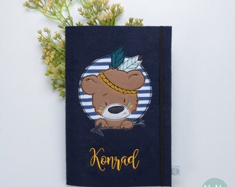 May include: A dark blue felt notebook cover with a brown bear wearing a feather headdress and a yellow headband. The bear is in a circle with blue and white stripes. The name Konrad is embroidered in yellow below the bear. The cover has a black elastic closure.