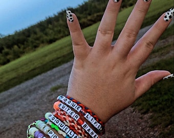 May include: A person's wrist with several colorful friendship bracelets. The bracelets have the words "Rogue Life", "Outer Banks", "Poguelandia", "Pogues", and "For Life" on them.