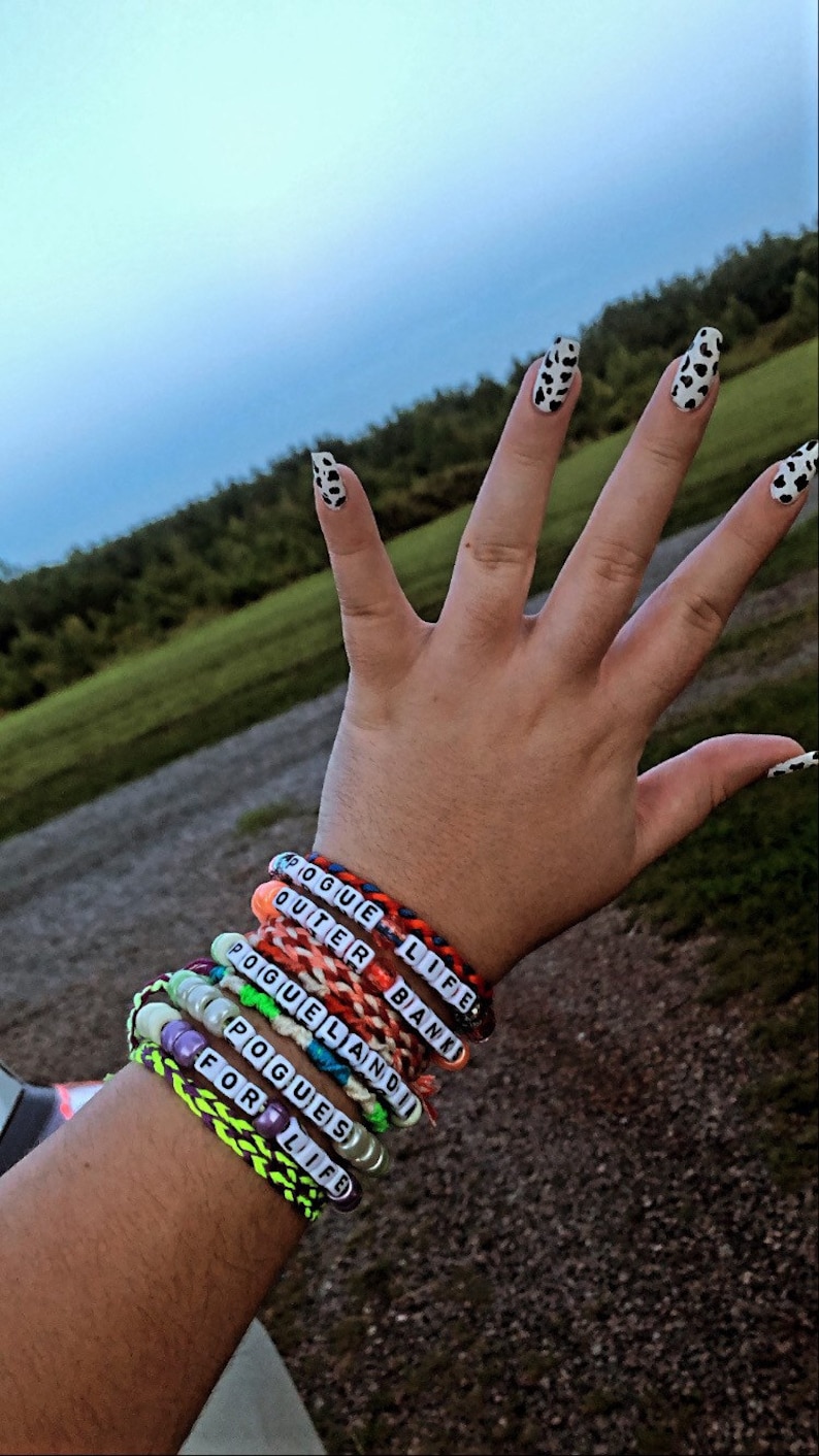 Puede incluir: Una mu&ntilde;eca con varios brazaletes de amistad de colores. Los brazaletes tienen las palabras "Rogue Life", "Outer Banks", "Poguelandia", "Pogues" y "For Life" en ellos.