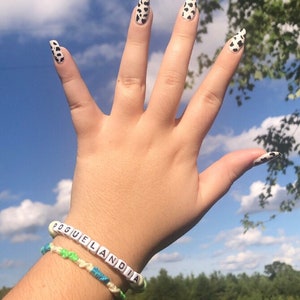 Puede incluir: Una mano con esmalte de u&ntilde;as de estampado de vaca y una pulsera que dice "Pogue Land". La mano est&aacute; levantada en el aire contra un cielo azul con nubes blancas.