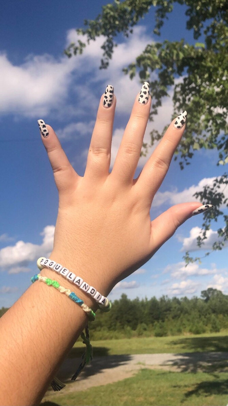 Puede incluir: Una mano con esmalte de u&ntilde;as de estampado de vaca y una pulsera que dice "Pogue Land". La mano est&aacute; levantada en el aire contra un cielo azul con nubes blancas.