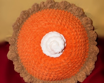 Puede incluir: Un cojín redondo, suave y tejido a ganchillo con forma de tarta de calabaza. El centro es de color naranja brillante con una superficie texturizada de hilo, rodeado por un borde de corteza marrón claro con volantes. Un copete blanco en el centro.