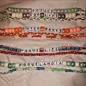 Puede incluir: Cuatro pulseras de cuentas con las palabras "Pogues for Life", "Outer Banks", "Pogue Life" y "Poguelandia" deletreadas en cuentas de letras blancas.