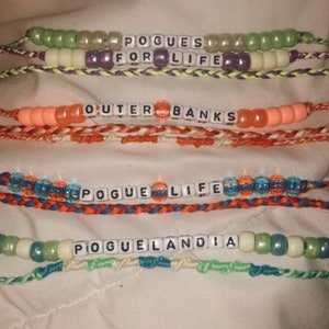 Puede incluir: Cuatro pulseras de amistad de colores con las palabras "Pogues for Life", "Outer Banks", "Pogue Life" y "Poguelandia" escritas en cuentas de letras blancas.