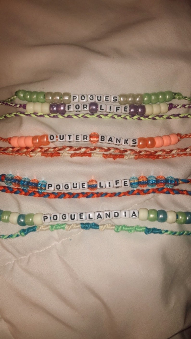 Puede incluir: Cuatro pulseras de amistad de colores con las palabras "Pogues for Life", "Outer Banks", "Pogue Life" y "Poguelandia" escritas en cuentas de letras blancas.
