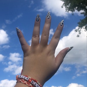 Puede incluir: Una mano con esmalte de u&ntilde;as de estampado de vaca blanco y negro contra un cielo azul con nubes blancas. La mano lleva una pulsera de amistad roja y blanca con las palabras "Outer Banks" escritas en cuentas blancas.