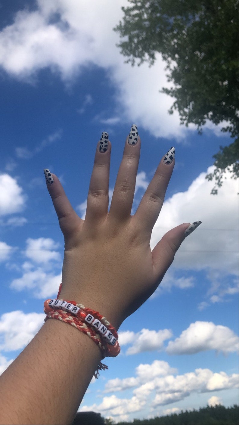 Puede incluir: Una mano con esmalte de u&ntilde;as de estampado de vaca blanco y negro contra un cielo azul con nubes blancas. La mano lleva una pulsera de amistad roja y blanca con las palabras "Outer Banks" escritas en cuentas blancas.