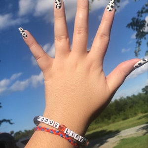 Puede incluir: Una mano con u&ntilde;as largas y blancas con un dise&ntilde;o de vaca en blanco y negro. La mano lleva una pulsera con las palabras "Pogue Life" en cuentas blancas.