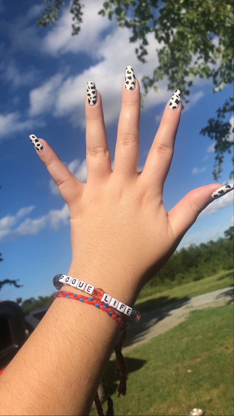 Puede incluir: Una mano con u&ntilde;as largas y blancas con un dise&ntilde;o de vaca en blanco y negro. La mano lleva una pulsera con las palabras "Pogue Life" en cuentas blancas.