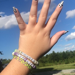 Puede incluir: Dos pulseras de cuentas con las palabras "Pogues for Life" en ellas. Las pulseras est&aacute;n en la mu&ntilde;eca de una persona con esmalte de u&ntilde;as de estampado de vaca en los dedos.