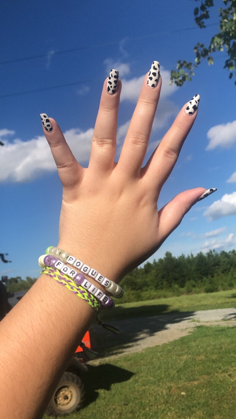 Puede incluir: Dos pulseras de cuentas con las palabras "Pogues for Life" en ellas. Las pulseras est&aacute;n en la mu&ntilde;eca de una persona con esmalte de u&ntilde;as de estampado de vaca en los dedos.