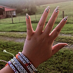 Puede incluir: Una mano con varios brazaletes de cuentas con las palabras "POGUE LIFE", "OUTER BANKS", "POGUELANDIA", "POGUES" y "FOR LIFE" en ellos.