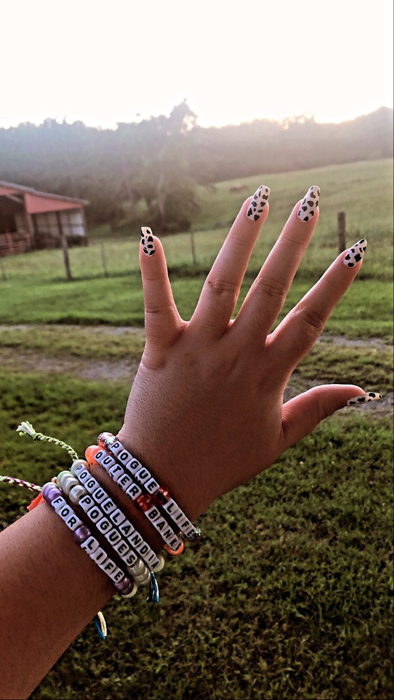 Puede incluir: Una mano con varios brazaletes de cuentas con las palabras "POGUE LIFE", "OUTER BANKS", "POGUELANDIA", "POGUES" y "FOR LIFE" en ellos.