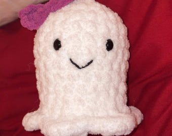 Puede incluir: Un fantasma de peluche tejido a ganchillo en color blanco con un acabado de hilo texturizado y esponjoso. El fantasma tiene dos pequeños ojos negros circulares y una sencilla sonrisa bordada en negro. Un pequeño lazo morado está en la parte superior.