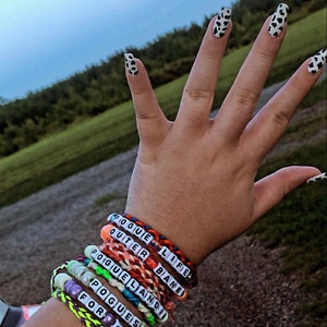 Puede incluir: Una mu&ntilde;eca con varios brazaletes de cuentas de colores. Los brazaletes tienen las palabras "Pogue Life", "Outer Banks", "Poguelandia", "Pogue's" y "For Life" en ellos. El texto "all bracelets" est&aacute; escrito debajo de los brazaletes.