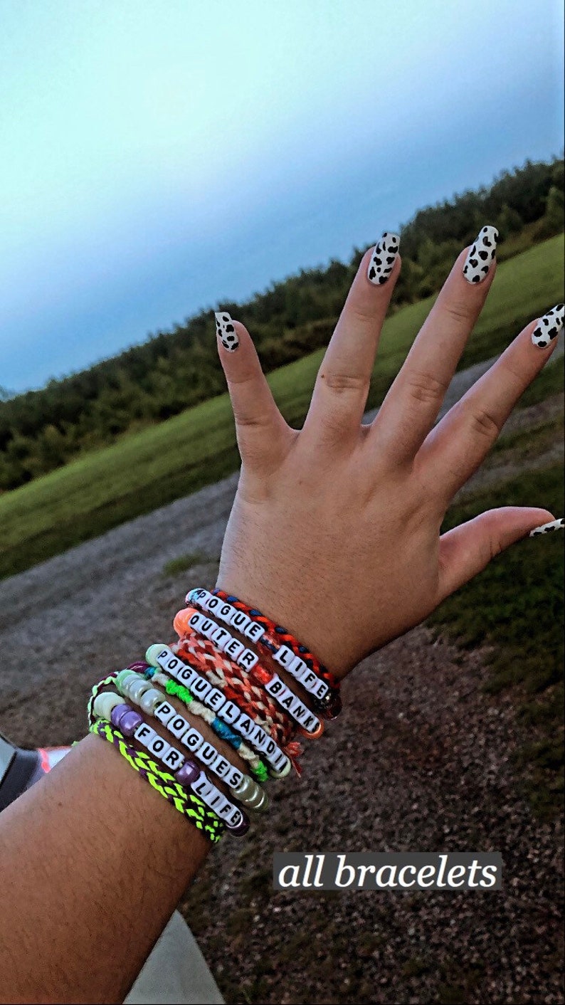 Puede incluir: Una mu&ntilde;eca con varios brazaletes de cuentas de colores. Los brazaletes tienen las palabras "Pogue Life", "Outer Banks", "Poguelandia", "Pogue's" y "For Life" en ellos. El texto "all bracelets" est&aacute; escrito debajo de los brazaletes.