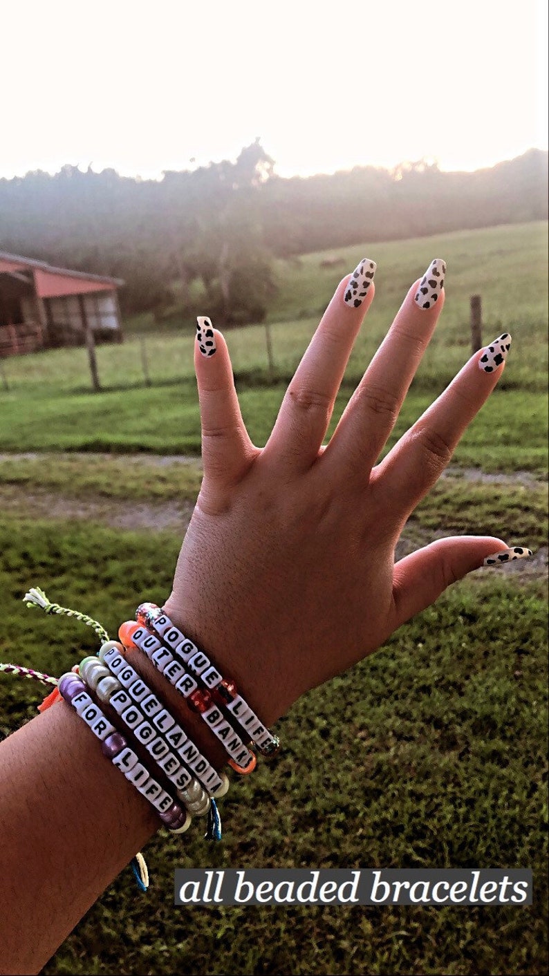 Puede incluir: Una mu&ntilde;eca con varios brazaletes de cuentas de colores. Los brazaletes tienen las palabras "POGUE LIFE", "OUTER BANKS", "POGUELANDIA", "POGUES" y "FOR LIFE" escritas en ellos.