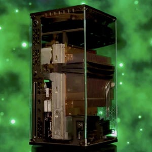 以下が含まれることがあります： 透明な長方形のディスプレイケースで、コンピューターの内部コンポーネントを紹介しています。コンポーネントには、マザーボード、ヒートシンク、その他の電子部品が含まれます。背景は、白い光るオーブのある緑色の星雲のようなデザインです。
