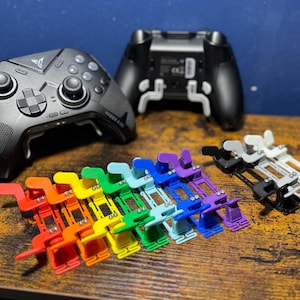 Puede incluir: Un conjunto de 12 accesorios de mando de juego de plástico de colores, dispuestos en un patrón de arcoíris. Los accesorios están diseñados para ser utilizados con un mando de juego para mejorar el agarre y el control. También se pueden ver dos mandos de juego negros en el fondo.