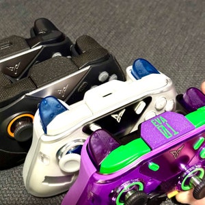Può includere: Tre controller per videogiochi di diversi colori. Il primo è nero con un logo argentato, il secondo è bianco con un pulsante blu e un logo verde, e il terzo è viola con un pulsante verde e un logo verde. Il controller viola ha il testo "EVA 01" su di esso.