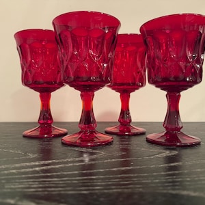 Può includere: Quattro calici da vino rosso rubino con un motivo a diamante sulla coppa e un gambo. I calici sono disposti su una superficie di legno scuro. I calici sono di vetro.