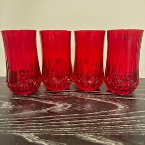 May include: Four red drinking glasses with a faceted base and a smooth, slightly curved body. The glasses are made of a transparent material and are arranged in a row on a dark wooden surface. The glasses are empty.