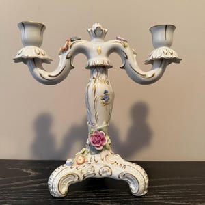 Puede incluir: Un candelabro de porcelana blanca con dos brazos, adornado con ribetes dorados y detalles florales. La base presenta un diseño detallado con una rosa rosa y otras flores coloridas. Los candelabros tienen un borde festoneado.