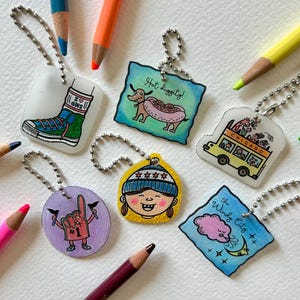May include: A collection of colorful keychains with Chicago-themed illustrations, including a hot dog, a school bus, and a person wearing a winter hat. The keychains are surrounded by colored pencils.