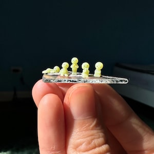 Peut inclure: Cinq figurines miniatures jaune pâle aux traits simples sont disposées en rangée sur un petit morceau de verre triangulaire transparent et texturé. Les figurines sont représentées dans diverses poses, allongées, assises ou debout.