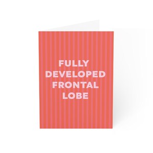 Puede incluir: Tarjeta de felicitación plegada con un diseño a rayas rojas y rosas. La parte delantera de la tarjeta muestra el texto "FULLY DEVELOPED FRONTAL LOBE" en rosa claro. El interior de la tarjeta es blanco.