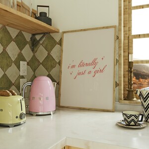 May include: A pink electric kettle with a silver handle and base sits on a white countertop next to a yellow toaster. A framed print with the text "i'm literally just a girl" hangs on the wall behind the appliances.
