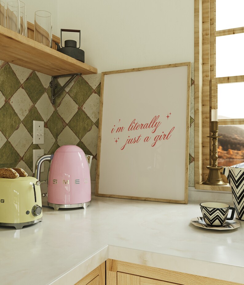 May include: A pink electric kettle with a silver handle and base sits on a white countertop next to a yellow toaster. A framed print with the text "i'm literally just a girl" hangs on the wall behind the appliances.