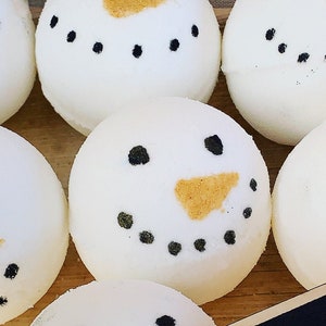 May include: A collection of white, spherical bath bombs designed as snowmen. Each bath bomb features black dot eyes and a mouth, with an orange triangle nose. The bath bombs are displayed on a wooden surface.