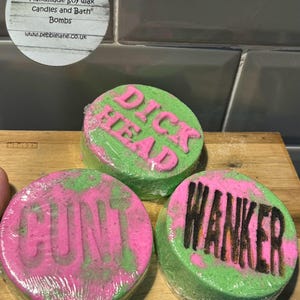 May include: Three round bath bombs with green and pink swirls. The bath bombs have the words "DICK HEAD", "CUNT", and "WANKER" printed on them in black.