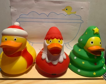 May include: Three rubber duck bath toys decorated for Christmas. The first duck is wearing a red and white Santa hat and a red scarf. The second duck is wearing a red Santa suit with a white trim. The third duck is shaped like a green Christmas tree with white and yellow stars.