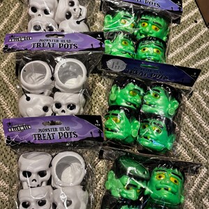 May include: Multiple packs of Halloween treat pots. Some are white skull-shaped, others are green Frankenstein monster heads. Each pack contains four pots and is labelled "Monster Head Treat Pots" with "Fantastic Halloween" branding.