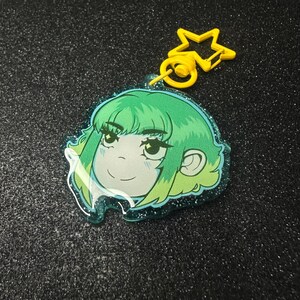 May include: A green-haired anime-style character keyring with a yellow star-shaped clasp. The character has a smiling expression and is outlined in a teal glittery border. The background is a dark, glittery surface.