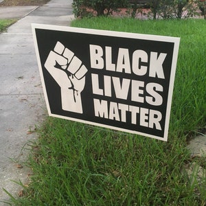 May include: A black and white sign with the text "BLACK LIVES MATTER" and a raised fist graphic. The sign is placed in a grassy area.