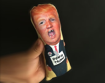 Peut inclure: Une poupée en peluche fantaisie représentant une caricature d'un homme aux cheveux orange et en costume. La poupée tient une pancarte avec l'inscription "PUNCH TRUMP". La poupée a également un bouton qui dit "I Putin".
