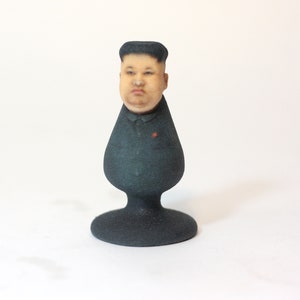 Puede incluir: Una figura impresa en 3D en negro y gris de la cabeza de un hombre con una expresión seria. La figura lleva una chaqueta de traje negra con una estrella roja en la solapa. La figura está montada sobre una base negra.