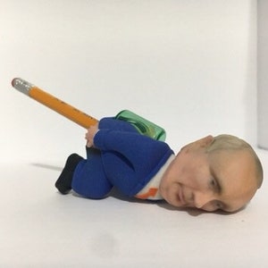 May include: A blue and orange figurine of a person lying on their back with a yellow pencil resting on their body. The figurine is wearing a blue suit and has a green object on their back.