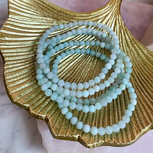 Può includere: Un set di quattro braccialetti di perline blu chiaro impilati su un piatto a forma di foglia dorata. I braccialetti sono fatti di perline rotonde e lisce.