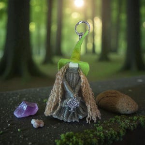 May include: A handcrafted fairy keyring with a green hat and long, flowing, light brown yarn hair. The fairy wears a grey dress and a silver tree of life pendant. A purple crystal and a small stone are nearby.