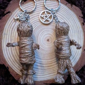 Peut inclure: Deux porte-cl&eacute;s faits &agrave; la main avec de petites figurines de poup&eacute;es envelopp&eacute;es faites de ficelle. Un porte-cl&eacute;s a un charme d'arbre, l'autre un charme de pentagramme. Les porte-cl&eacute;s sont pr&eacute;sent&eacute;s sur une tranche de bois.