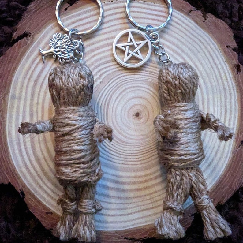 Peut inclure: Deux porte-cl&eacute;s faits &agrave; la main avec de petites figurines de poup&eacute;es envelopp&eacute;es faites de ficelle. Un porte-cl&eacute;s a un charme d'arbre, l'autre un charme de pentagramme. Les porte-cl&eacute;s sont pr&eacute;sent&eacute;s sur une tranche de bois.